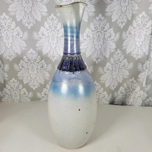 May include: A tall, ceramic vase with a white, blue, and navy color scheme. The vase has a flared top and a textured, ribbed neck. The body of the vase is a rounded, elongated shape.