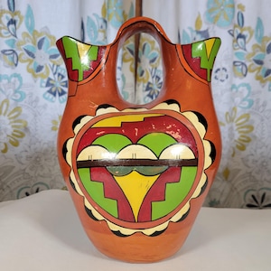 May include: A hand-painted orange ceramic pitcher with a geometric design in red, green, yellow, and black. The pitcher has two spouts and a handle.