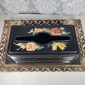 May include: A rectangular black tissue box cover featuring painted floral motifs in shades of yellow, orange, and green, resting on a decorative gold-toned metal tray with an intricate filigree border and a mirrored base.