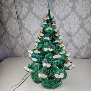 May include: A green ceramic Christmas tree with a glossy finish, featuring white painted snow on the branches and numerous small, multi-colored plastic light bulbs inserted throughout the structure, topped with a single blue star light.