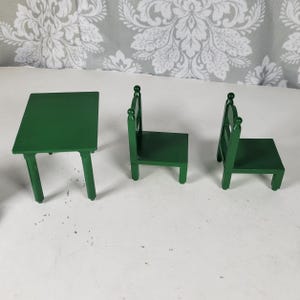 Puede incluir: Un juego de mesa y dos sillas de madera verde en miniatura. La mesa tiene cuatro patas y una tapa rectangular. Las sillas tienen un respaldo con una parte superior decorativa. El conjunto est&aacute; pintado en color verde liso.