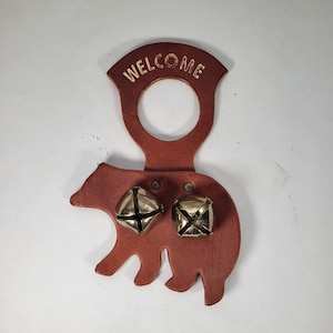 May include: A brown leather bear-shaped welcome sign with two brass bells. The word "WELCOME" is embossed at the top. The sign is designed to hang and has a rustic, handcrafted appearance.
