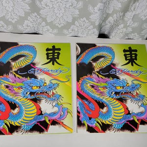 May include: Two colorful Ed Hardy art prints featuring a dragon design. The dragons are blue, yellow, red, and pink, set against a lime green and black background. The prints include the text "東" and "Ed Hardy" in a stylized font.
