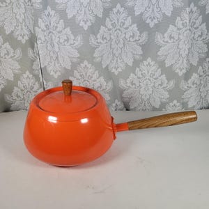 May include: An orange enamel saucepan with a wooden handle and lid. The lid has a wooden knob. The saucepan is round with a long handle. The handle and knob are made of wood. The background is a gray and white floral pattern.
