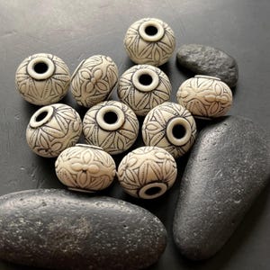 May include: Ivory-colored, round beads with black etched floral patterns and a central hole. The beads are arranged with dark gray stones on a dark surface. The beads have a vintage or bohemian aesthetic.