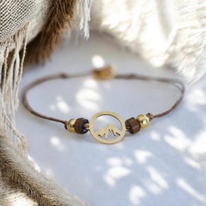 Könnte beinhalten: Ein Armband mit einem goldfarbenen Bergketten-Anhänger. Der Anhänger ist ein kreisförmiges Design mit einer Bergsilhouette. Das Armband hat braune und beige Kordeln mit Holz- und goldfarbenen Perlen. Das Armband wird auf einer weißen Oberfläche präsentiert.