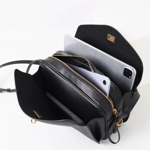 May include: Black crossbody bag with gold-toned hardware, open to reveal a tablet and a smartphone. The bag has a rectangular shape with a flap closure and a long, adjustable strap. The interior is lined with a light-colored fabric.