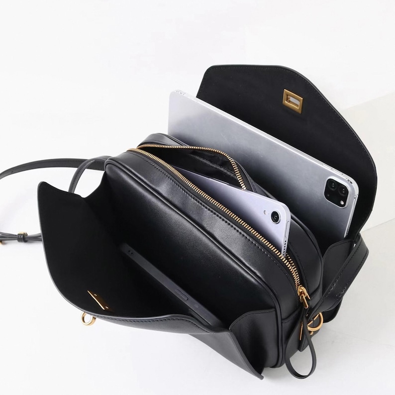 May include: Black crossbody bag with gold-toned hardware, open to reveal a tablet and a smartphone. The bag has a rectangular shape with a flap closure and a long, adjustable strap. The interior is lined with a light-colored fabric.