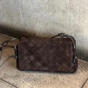 May include: A dark brown rectangular shoulder bag featuring a woven leather exterior pattern. The bag includes a matching dark brown leather shoulder strap that is secured on each side with a simple knot detail, providing a structured appearance.