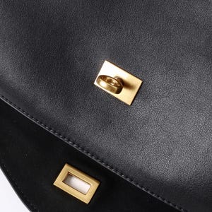 May include: Close-up of a black leather handbag with gold-colored hardware. The bag features a rectangular clasp and a circular ring closure. The leather has a textured appearance.