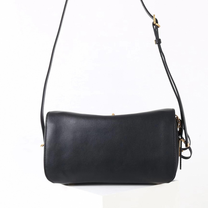 May include: Black crossbody bag with a long, adjustable strap. The rectangular bag has a curved top and a gold-colored clasp. The strap is thin and black, and the bag appears to be made of leather or a similar material. The bag is shown against a white background.
