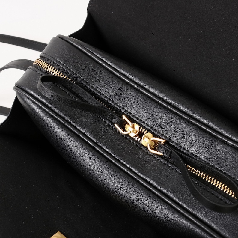 May include: Black leather crossbody bag with a gold zipper and a black leather pull. The bag has a rectangular shape and a flap closure. The interior of the bag is black.