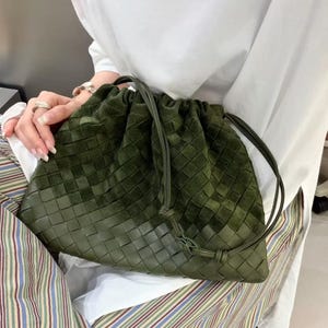 Könnte beinhalten: Eine dunkel olivgrüne Umhängetasche aus geflochtenem Leder mit einer weichen, wildlederartigen Textur. Die Tasche verfügt über einen Kordelzugverschluss an der Oberseite, der einen gerafften Effekt erzeugt, sowie einen dünnen, passenden Schulterriemen.