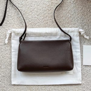 May include: A dark brown rectangular crossbody bag with a pebbled texture and a thin shoulder strap. The bag features a simple, clean design with a small, subtle logo embossed on the front center. It is presented against a plain white fabric pouch.