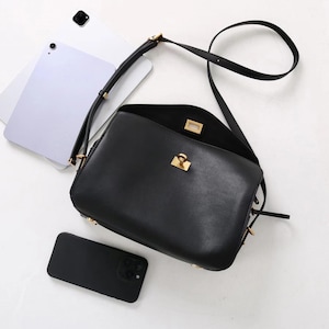 May include: Black leather crossbody bag with a gold-tone clasp and hardware. The bag is open, revealing the interior. A black smartphone and a silver tablet are next to the bag. The bag has a long, adjustable strap.