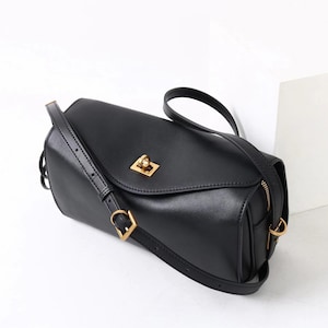 May include: Black rectangular crossbody bag with a gold-colored clasp and hardware. The bag has a long, adjustable strap and a zipper closure. The bag is made of a smooth, leather-like material.