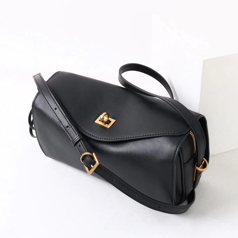 May include: Black rectangular crossbody bag with a gold-colored clasp and hardware. The bag has a long, adjustable strap and a zipper closure. The bag is made of a smooth, leather-like material.