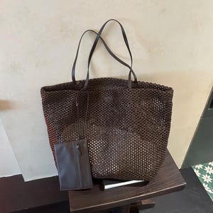 Può includere: Una borsa tote marrone intrecciata con manici coordinati e una piccola pochette rettangolare. La borsa presenta un design a trama aperta. La pochette ha una chiusura a zip. La borsa è su una superficie di legno.