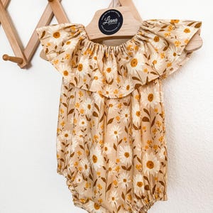 May include: A beige romper featuring a floral pattern of white daisies and small yellow flowers. The garment has a wide, ruffled collar and elasticized leg openings. It is displayed on a wooden hanger with a circular Liana Handmade logo.