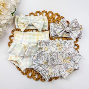 May include: Baby clothing set featuring two pairs of ruffled bloomers and matching headbands. One set has a pastel plaid pattern, and the other has a floral print with bunnies and eggs. The headbands have large bows.