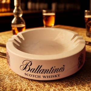 Puede incluir: Un cenicero rosa claro con el logotipo de Ballantine's Scotch Whisky. El cenicero tiene cuatro ranuras para cigarrillos y está rodeado de vasos de líquido ámbar. Una botella de licor está en el fondo.