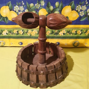 Puede incluir: Un cascanueces de madera con una base de barril y dos brazos para cascar nueces. El cascanueces está hecho de madera marrón oscura y tiene un aspecto rústico.