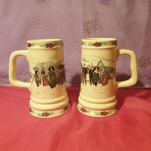 Puede incluir: Dos jarras de cerveza de cerámica blanca con un diseño floral alrededor de la parte superior e inferior. Cada jarra presenta una ilustración colorida de personas en una escena nevada.