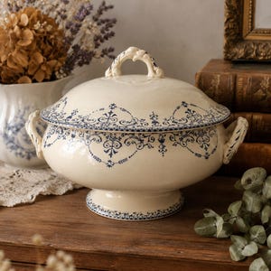 Peut inclure: Une soupière en céramique de couleur crème avec un couvercle assorti, ornée de motifs floraux et de volutes bleus complexes. La soupière possède deux anses latérales, une base sur pied et une poignée décorative sur le couvercle.