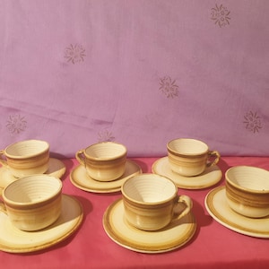 May include: Set of six brown and white ceramic teacups and saucers. The cups and saucers have a simple design with a brown band around the rim and a white interior.