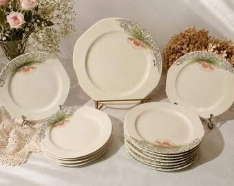May include: A set of cream-colored ceramic plates featuring an octagonal shape and a decorative pattern of a pink flower with green leaves and a fine black speckled border. The collection includes one large serving plate and several smaller plates.