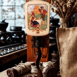 Op de afbeelding: Oude koffiemolen met een crèmekleurige keramische container met een kleurrijke illustratie van mensen in traditionele kleding. De molen heeft een houten basis, metalen onderdelen en een houten handvat. Koffiebonen liggen op het jute oppervlak.