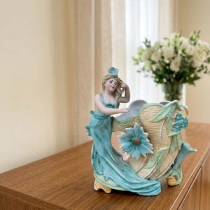 Pode incluir: Um vaso com estatueta de porcelana apresentando uma figura num vestido fluido azul-petróleo com uma flor a combinar no cabelo. A figura apoia-se num recipiente cor de creme em forma de concha decorado com uma grande flor azul e folhas.