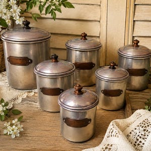 May include: A set of five cylindrical metal canisters with removable lids and dark copper-toned labels. Each canister features a metallic silver finish, a small round knob on the lid, and a rectangular plaque on the front secured by two rivets.