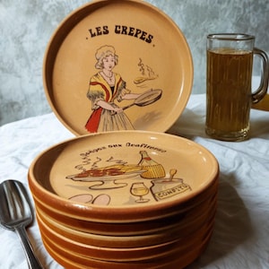 Peut inclure: Une pile d'assiettes en céramique beige avec un bord marron, présentant des illustrations de crêpes et de nourriture. Une assiette montre une femme cuisinant des crêpes, et une autre porte l'inscription "Bonne aux Confitures". Une chope de liquide et une cuillère sont également visibles.