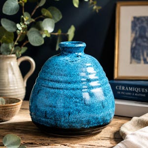 Peut inclure: Un vase en céramique rond et bombé avec un glaçage bleu vif moucheté et une base sombre. Le vase présente un col étroit et une série de stries horizontales entourant le corps, créant une surface texturée avec une finition brillante.