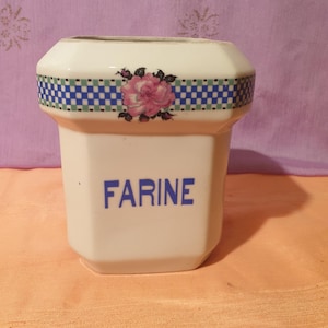 May include: White ceramic flour container with a blue and green checkered band and a pink rose design. The container has the word "FARINE" written in blue letters.