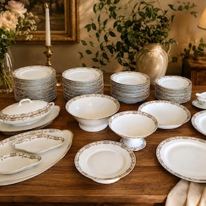 Peut inclure: Un grand service de table en porcelaine blanche orné d'un délicat motif floral doré et gris sur les bords. La collection comprend des assiettes empilées, une soupière avec couvercle, un bol sur pied, des plats de service ovales et des coupelles.