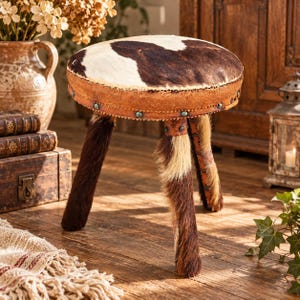 May include: A round stool featuring a cowhide seat with brown and white patches. The side of the seat is wrapped in textured brown leather secured with metal studs. The stool stands on three legs covered in long, coarse brown and white fur.