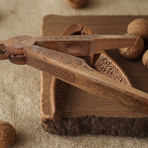 May include: A wooden nutcracker featuring a hinged design with intricate geometric cross-hatch carvings along the handles. The tool is positioned with several whole walnuts nearby, showcasing the natural brown wood grain and detailed craftsmanship.