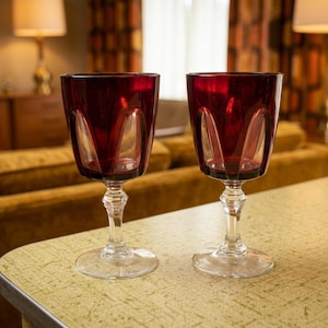 Op de afbeelding: Twee robijnrode wijnglazen met heldere, sierlijke stelen en voetjes. De glazen hebben een gesneden ontwerp op de kom. De glazen staan op een gele tafel met een retro-ontwerp. De achtergrond omvat een bank en gordijnen.