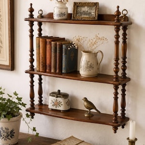 May include: A three-tiered dark wood wall shelf with turned spindle supports. The top shelf holds a small white floral vase and a framed picture. The middle shelf displays five vintage books and a white pitcher. The bottom shelf holds a floral box and a brass bird figurine.