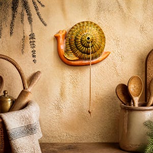 May include: A ceramic wall-mounted snail sculpture featuring a glossy, golden-brown spiral shell and a smooth orange body. A thin, light-colored string hangs vertically from the center of the shell, ending in a small knot at the bottom of the piece.
