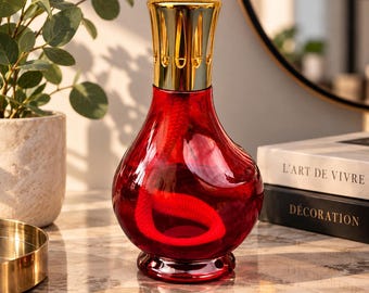 Könnte beinhalten: Eine rote Duftlampe aus Glas mit bauchigem Körper und einer goldfarbenen Metallkrone. Ein weißer Docht ist im Inneren des transparenten roten Glasgefäßes sichtbar. Die Lampe steht auf einer Marmoroberfläche neben Büchern mit Titeln.