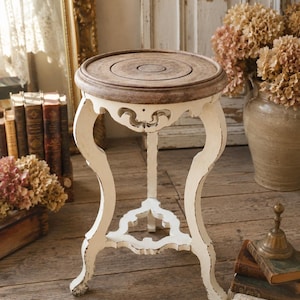 May include: A round side table featuring a weathered brown wooden top and a distressed off-white base with curved legs. The table includes a decorative lower shelf connecting the three legs, showcasing a worn finish on the painted wooden structure.