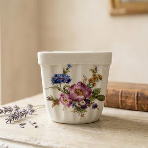 Peut inclure: Un pot en céramique blanche avec un extérieur cannelé et un motif floral composé de bleuets bleus, de roses roses et de petites fleurs jaunes. Le récipient présente un bord lisse et droit ainsi qu'un corps fuselé avec une illustration botanique.