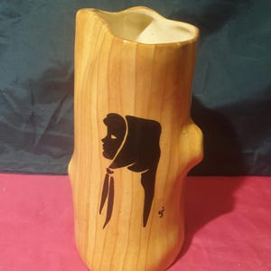 May include: A ceramic vase shaped like a tree trunk with a silhouette of a woman's head wearing a headscarf. The vase is painted in shades of brown and black.