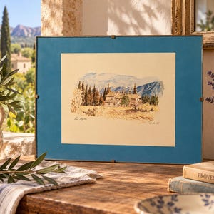 Könnte beinhalten: Ein rechteckiges gerahmtes Aquarellbild, das eine ländliche Landschaft mit Steingebäuden, Zypressen und fernen Bergen zeigt. Das Kunstwerk enthält den handschriftlichen Text Les Alpilles sowie eine Signatur mit dem Datum 85 auf cremefarbenem Grund.