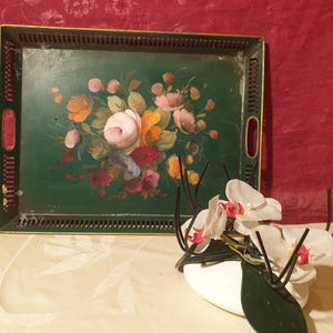 May include: A green metal tray with a floral design painted on it. The tray has a black metal frame and a decorative border. The tray is sitting on a white surface with a white flower arrangement.