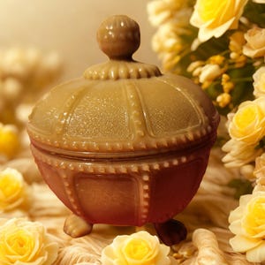 Puede incluir: Un joyero vintage con tapa, en tonos beige y rosa. El joyero tiene forma redondeada, superficie texturizada y detalles decorativos. Se apoya sobre tres pequeños pies y tiene un asa en forma de pomo. Rosas amarillas en el fondo.