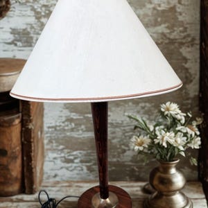 May include: A vintage table lamp with a white lampshade and a dark wood base. The lampshade has a brown trim. The lamp sits on a wooden surface next to a vase of white flowers. A black power cord is visible.